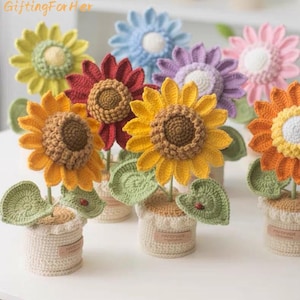 May include: A collection of handmade crochet sunflowers in various colors, including yellow, red, and orange. Each flower has a detailed center and green leaves, potted in small, off-white crochet pots. The text "GiftingForHer" is in the top left corner.