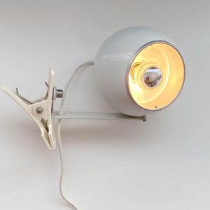 Puede incluir: Una lámpara de pinza esférica blanca con acabado brillante y borde interior plateado. La luz está unida a un mecanismo de sujeción de resorte blanco con una bisagra de metal y un cable de alimentación blanco que sale de la parte posterior.