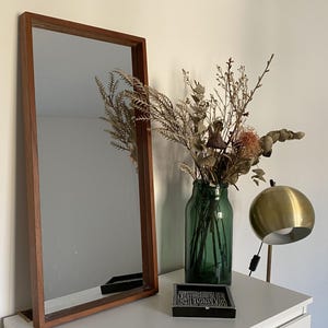 May include: A rectangular mirror with a brown wooden frame, a green glass vase containing dried botanical stems, a small square black and white patterned tray, and a spherical brass lamp with a white interior shade and a black power cord.