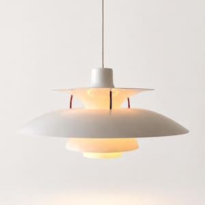 May include: A white pendant light fixture featuring a multi-tiered shade design with three distinct circular layers. The structure is held together by thin vertical metal connectors, and the lamp emits a soft, warm glow from the center.