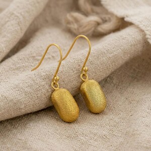 May include: A pair of gold-colored earrings with a textured, bean-shaped design. The earrings hang from delicate gold hooks and are displayed on a neutral-toned fabric background. The earrings have a matte finish and a subtle, organic shape.