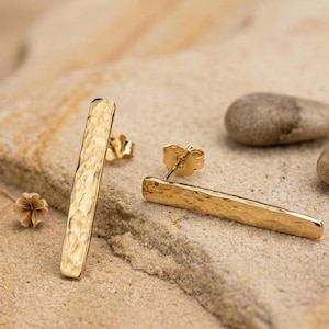 Puede incluir: Un par de pendientes de barra dorados con textura martillada. Los pendientes se exhiben sobre una superficie arenosa con pequeñas piedras y una flor seca. Los pendientes tienen cierre de poste.
