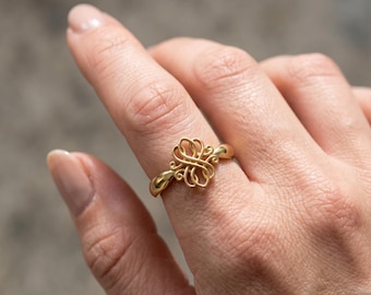 May include: A gold ring with an intricate, swirling design. The ring's focal point is a detailed, openwork pattern, and the band is smooth and polished. The ring is shown on a finger, highlighting its delicate craftsmanship and elegant aesthetic.