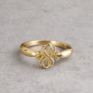 Puede incluir: Un anillo dorado con un diseño ornamental y simétrico en el centro. La banda del anillo es lisa y redondeada, lo que conduce a un patrón detallado y arremolinado en el centro. El anillo está sobre un fondo gris neutro.