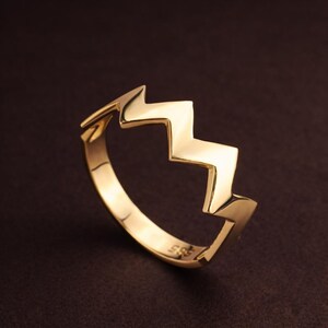 Può includere: Un anello color oro con un design a zig-zag. L'anello ha una finitura liscia e lucida e un design moderno. L'anello è mostrato su uno sfondo marrone scuro.