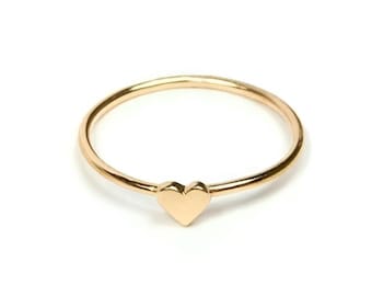 May include: A delicate gold ring featuring a small, solid heart charm. The ring has a thin band and a polished finish, set against a white background. The heart is centered on the ring, creating a simple yet elegant design.
