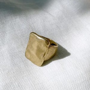 May include: A gold-coloured ring with a textured, rectangular face. The ring has a thick band and a slightly irregular shape, giving it a unique, handcrafted appearance. The ring is set against a white, textured background.