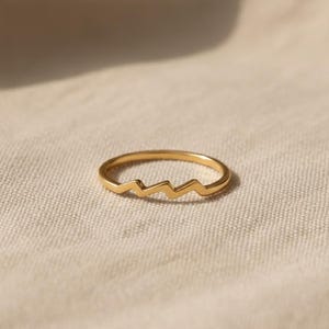 Könnte beinhalten: Ein zarter goldener Ring mit Zickzack-Design. Der Ring ist dünn und hat eine polierte Oberfläche, die das Licht reflektiert. Der Ring ist vor einem neutralen Hintergrund platziert, was seine schlichte Eleganz unterstreicht. Ein minimalistisches Schmuckstück.