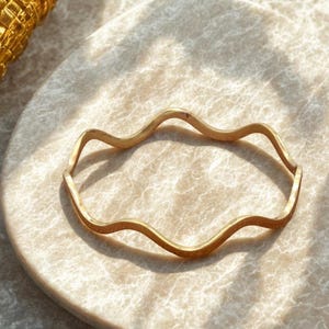 May include: A gold-colored bangle bracelet with a wavy, organic design. The bracelet has a textured surface and is set against a neutral background. The bracelet's unique shape and color make it a striking piece of jewelry.