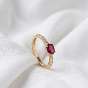 Peut inclure: Une bague en or ornée d'un rubis ovale et de petites pierres d'ornement. La monture est ornée de minuscules pierres claires. La bague est posée sur un fond blanc texturé.