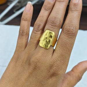 May include: A gold-colored ring with a rectangular face and a textured surface. The ring is worn on a hand, showcasing its design and craftsmanship. The ring's band is a simple, solid design.