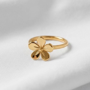 Può includere: Un anello dorato con un design floreale. L'anello ha una fascia sottile e un fiore a cinque petali. L'anello è posto su uno sfondo bianco, evidenziando la sua finitura metallica lucida. Il design dell'anello è semplice ed elegante.
