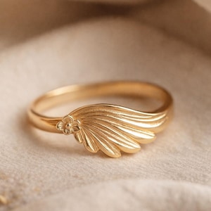 Op de afbeelding: Een goudkleurige ring met een bloemaccent en een vleugelachtig ontwerp. De ring heeft een klein bloemdetail en een reeks gebogen lijnen die de vleugelvorm creëren. De ring is waarschijnlijk gemaakt van verguld metaal.