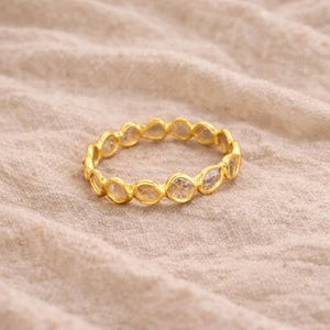 May include: A gold ring featuring a continuous line of clear, oval-cut gemstones. The ring is presented on a beige fabric, accentuating its warm gold hue and the brilliance of the stones. The design is delicate and refined.