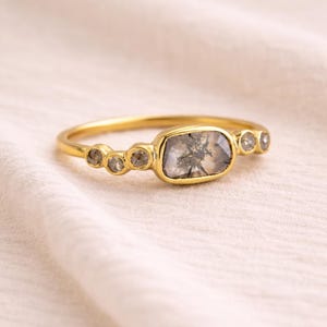 May include: A gold ring featuring a rectangular, gray-toned gemstone with internal patterns, flanked by three smaller, round, gray stones on each side. The ring is set against a soft, off-white background.
