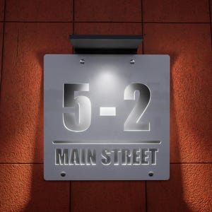 以下が含まれることがあります： 赤いレンガの壁を背景に、数字の「5-2」と「MAIN STREET」の文字が照らされた銀色の住所表示。看板は小さなネジで取り付けられており、上部に小さな照明器具が付いています。