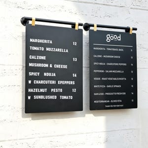 May include: Two rectangular black menu boards with white lettering, suspended from black metal bars. The left board lists pizza options like Margherita and Calzone with prices. The right board features the text The Good Life and various pizza items.