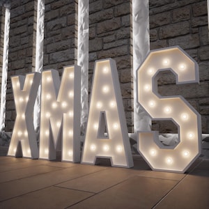 May include: White marquee letters spelling "XMAS" with warm white lights. The letters are standing on a wooden surface, with a brick wall and decorative columns in the background. Festive holiday decor.