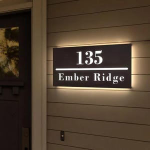 Puede incluir: Un letrero de dirección rectangular con el número "135" en blanco, encima de las palabras "Ember Ridge". El letrero es oscuro con una línea horizontal blanca que separa los números y el texto. El letrero está iluminado con una luz cálida.