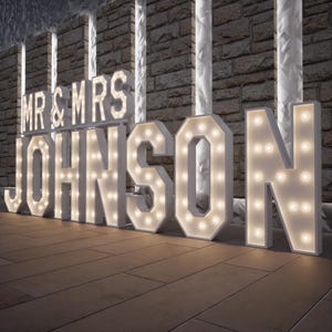 May include: Large white marquee letters spelling "MR & MRS JOHNSON" with warm white lights. The letters are on a wooden floor in front of a brick wall. Suitable for wedding or event decor.