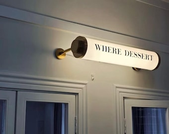May include: A horizontal cylindrical wall light fixture with a white frosted glass shade and dark metal end caps. The text WHERE DESSERT is printed in black capital letters on the front of the illuminated shade, which is mounted to the wall.