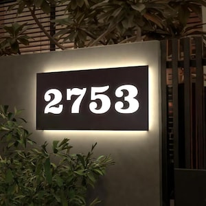 May include: A rectangular black address sign with the number "2753" in white, illuminated by a warm light. The sign is mounted on a gray wall, with greenery in the foreground and a dark background.