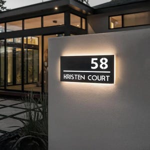 Puede incluir: Un letrero rectangular de metal negro para casa con iluminación de fondo. El letrero muestra el número 58 en una fuente grande sobre una línea horizontal, con el texto KRISTEN COURT en una fuente más pequeña debajo, montado en una pared clara.