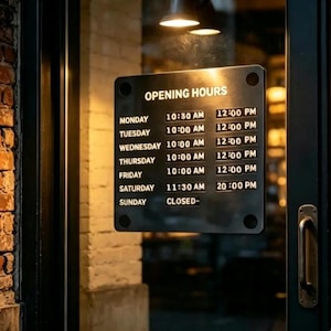 Puede incluir: Un letrero rectangular negro montado en una puerta de cristal que muestra el horario de apertura en texto blanco. El horario enumera de lunes a domingo con horas específicas, incluyendo de 10:30 a 12:00 el lunes y de 11:30 a 20:00 el sábado, domingo es CLOSED.