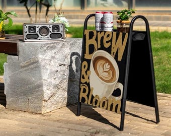 Puede incluir: Escena al aire libre con un letrero negro en forma de A que muestra "Brew & Bloom" y una ilustración de taza de café. Dos latas de cerveza encima. Un altavoz plateado sobre un bloque de piedra. Vegetación y un edificio al fondo.