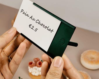 Puede incluir: Un soporte para letreros de metal verde oscuro con un marco rectangular y un pequeño detalle hexagonal. Una tarjeta rectangular blanca está insertada, con el texto manuscrito negro Pain Au Chocolat y €2.5.