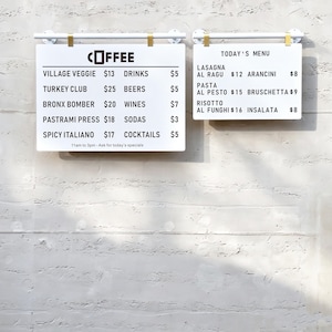 May include: Two white rectangular menu boards with black text hanging from white and gold metal bars. The left board lists coffee and food items like Village Veggie and Turkey Club with prices. The right board displays Today's Menu with Italian dishes.