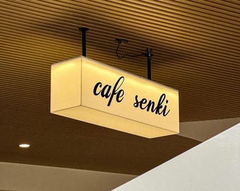 May include: A rectangular, illuminated sign with the words "cafe senki" in black script hangs from the ceiling. The sign has a soft, glowing appearance, and the background is a wooden ceiling.