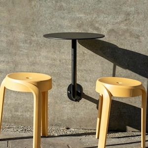 Puede incluir: Una mesa redonda negra montada en la pared con un solo soporte, junto con dos taburetes amarillos. Los taburetes tienen patas curvas y un asiento circular. La mesa está montada en una pared de hormigón gris, con sombras.