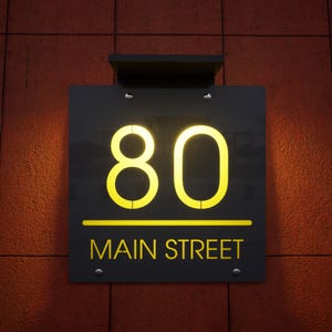 以下が含まれることがあります： 黒い長方形の住所看板。明るい黄色で「80」の数字が書かれ、その下に「MAIN STREET」の文字。赤いレンガの壁に取り付けられています。数字とテキストは照らされています。