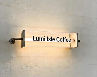 Puede incluir: Un letrero rectangular iluminado con el texto "Lumi Isle Coffee" en una fuente moderna. El letrero está encerrado en un marco de metal oscuro y montado en una pared texturizada de color claro. El letrero tiene un brillo cálido y acogedor.
