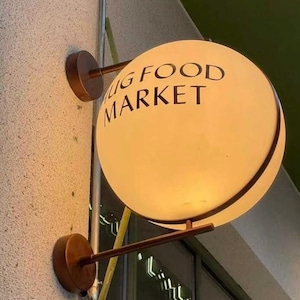 Könnte beinhalten: Ein rundes, beleuchtetes Schild für einen Lebensmittelmarkt. Das Schild ist eine leuchtende Kugel mit den Worten "BIG FOOD MARKET" in schwarzer Schrift. Es ist an einer Wand mit bronzefarbener Hardware befestigt.