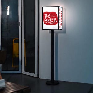 May include: A black, rectangular signpost with a white illuminated sign. The sign displays red text that reads "The Belega" and "Uncle Boggy's". The sign is mounted on a black pole and base, standing in front of a glass door.