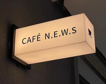 May include: Illuminated rectangular sign for a cafe, mounted on a dark wall. The sign reads "CAFÉ N.E.W.S" in black letters, with a small coffee cup icon on the right side. The sign is white and emits a soft glow.