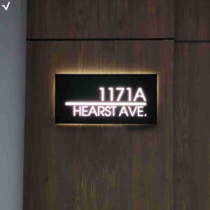 May include: A rectangular black address sign with backlit white lettering displaying 1171A HEARST AVE. The sign features a horizontal line separating the numbers from the street name, mounted on a vertical wood-grain panel with warm ambient lighting.