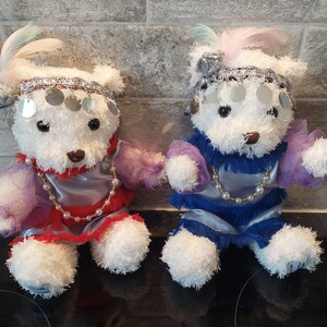 May include: Two white teddy bears in vibrant costumes. One is in red and silver, the other in blue and silver. Both have beaded necklaces, headbands with silver discs, and feather details.