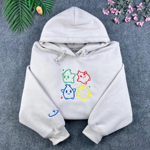 May include: A light gray hoodie with a drawstring hood and long sleeves. The front features four colorful cartoon star characters. A small blue embroidered design is on the left sleeve. The hoodie is made of a soft material.