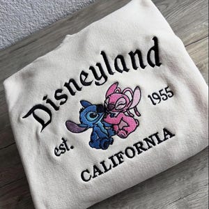 May include: Cream sweatshirt featuring black embroidered text reading "Disneyland", "est. 1955 CALIFORNIA", and a cartoon character design in blue and pink. The garment is folded on a wooden surface.