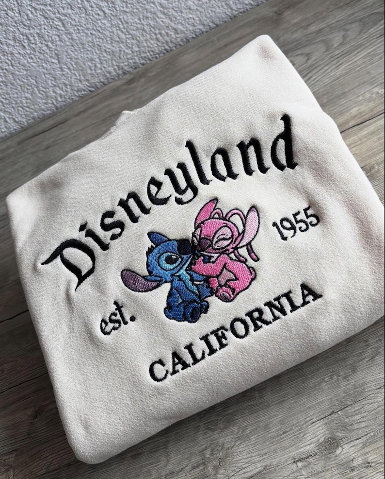 May include: Cream sweatshirt featuring black embroidered text reading "Disneyland", "est. 1955 CALIFORNIA", and a cartoon character design in blue and pink. The garment is folded on a wooden surface.