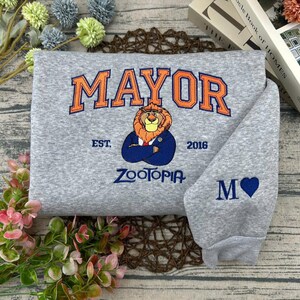 Puede incluir: Una sudadera gris con la palabra "MAYOR" en naranja sobre un le&oacute;n bordado con un traje azul. El texto "EST. 2016" y "ZOOTOPIA" est&aacute;n debajo del le&oacute;n. La manga tiene una "M" y un coraz&oacute;n azul.