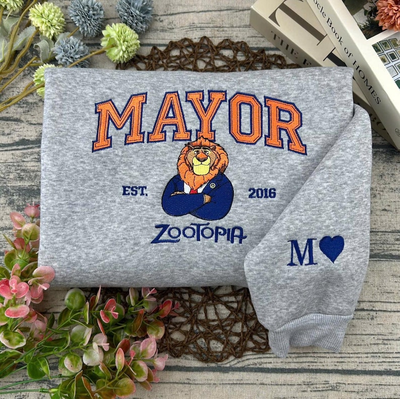 Puede incluir: Una sudadera gris con la palabra "MAYOR" en naranja sobre un le&oacute;n bordado con un traje azul. El texto "EST. 2016" y "ZOOTOPIA" est&aacute;n debajo del le&oacute;n. La manga tiene una "M" y un coraz&oacute;n azul.