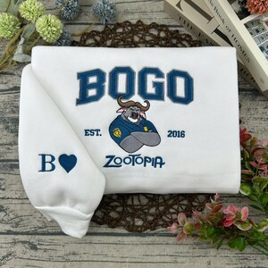 Puede incluir: Sudadera blanca con la palabra "BOGO" en azul y una ilustraci&oacute;n de un personaje de Zootopia. La manga muestra una "B" y un coraz&oacute;n azul. La sudadera tambi&eacute;n tiene el texto "EST. 2016" y "ZOOTOPIA".