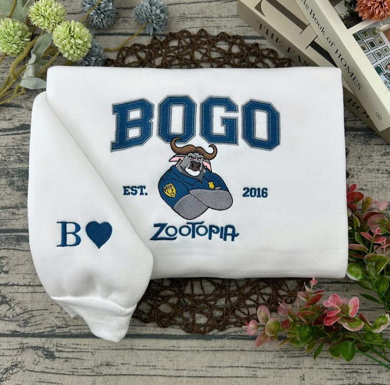 Puede incluir: Sudadera blanca con la palabra "BOGO" en azul y una ilustraci&oacute;n de un personaje de Zootopia. La manga muestra una "B" y un coraz&oacute;n azul. La sudadera tambi&eacute;n tiene el texto "EST. 2016" y "ZOOTOPIA".