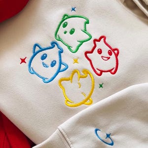 May include: Cream-colored hoodie with embroidered cartoon characters in blue, green, red, and yellow. Each character has a unique shape and expression. The design includes small star accents in various colors. The sleeve cuff features a small blue embroidered design.