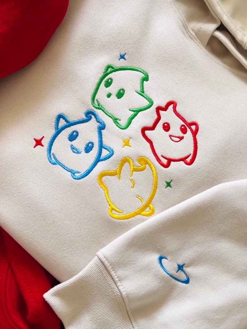 May include: Cream-colored hoodie with embroidered cartoon characters in blue, green, red, and yellow. Each character has a unique shape and expression. The design includes small star accents in various colors. The sleeve cuff features a small blue embroidered design.