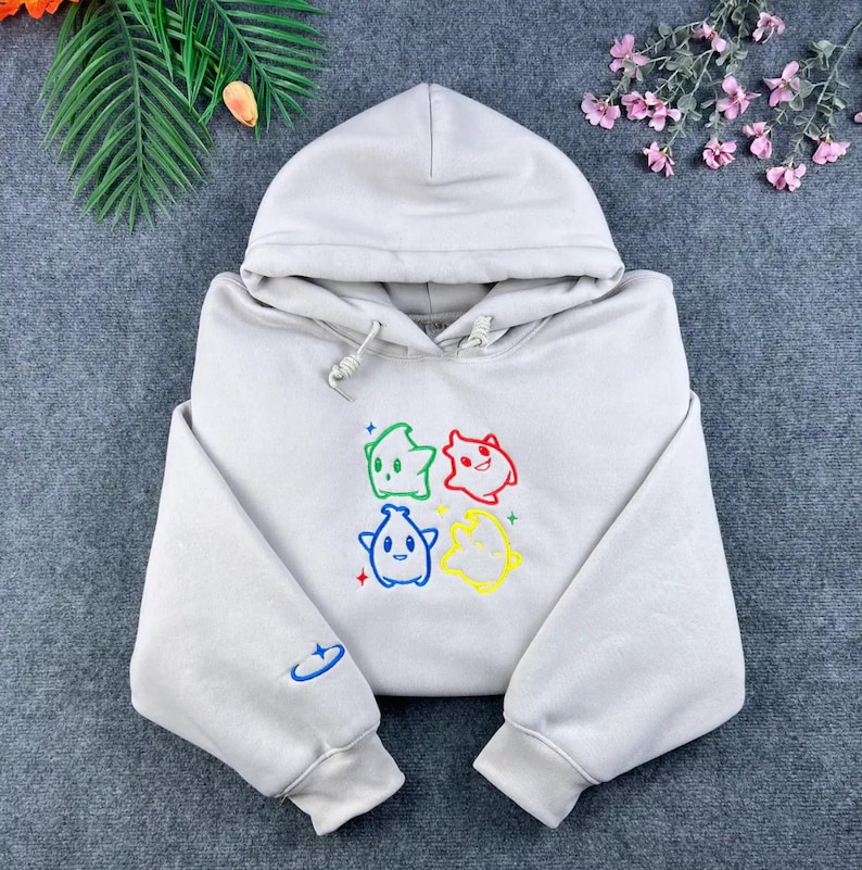 May include: A light gray hoodie with a drawstring hood and long sleeves. The front features four colorful cartoon star characters. A small blue embroidered design is on the left sleeve. The hoodie is made of a soft material.
